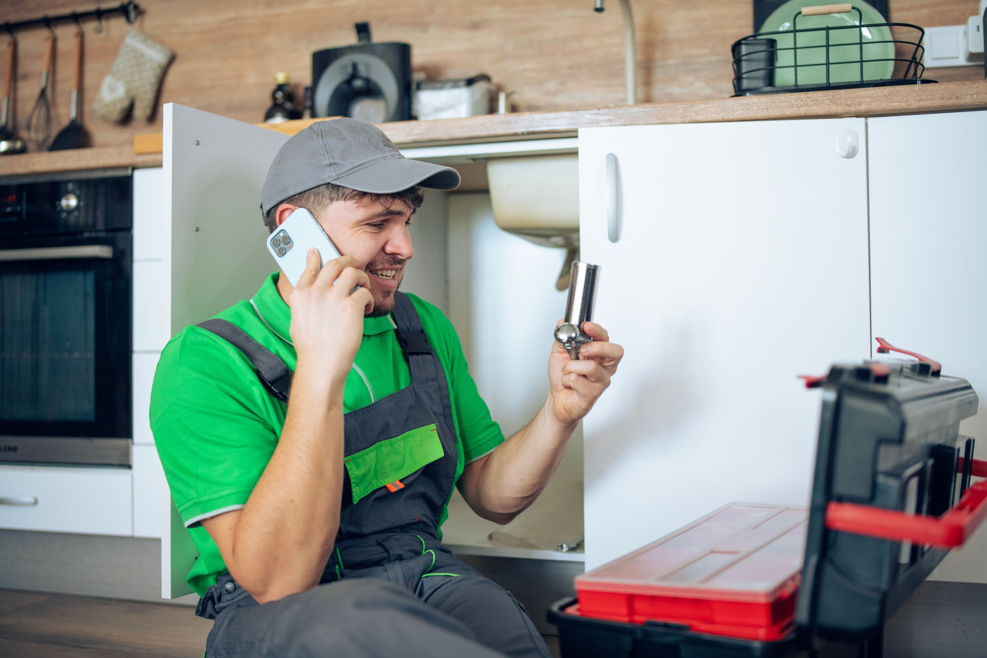 Plumber handling a phone call while fixing a kitchen sink pipe